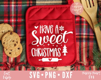 May include: Red oven mitt with the text "Have a Sweet Christmas" in white script. The mitt features a snowflake, Christmas tree, and rolling pin design. Chocolate chip cookies are on the left.