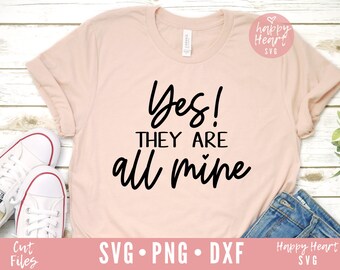 Könnte beinhalten: Ein rosa T-Shirt mit dem Text "Ja! Sie gehören alle mir" in schwarzer Schrift. Das Shirt liegt flach auf einer weißen Holzoberfläche mit einem Paar weißen Turnschuhen und einer blauen Jeans.