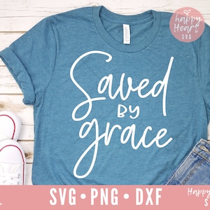 Puede incluir: Una camiseta azul turquesa con el texto "Saved by grace" en letra blanca. La camiseta est&aacute; puesta sobre una superficie de madera blanca con un par de zapatillas blancas y un par de pantalones vaqueros azules.