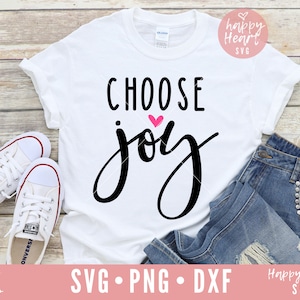 May include: White t-shirt with black text that says "CHOOSE joy" with a pink heart above the word "joy". The t-shirt is laying on a wooden surface with a pair of white sneakers and blue jeans.
