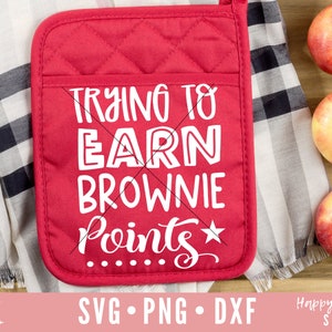 May include: A red quilted oven mitt with the text "TRYING TO EARN BROWNIE POINTS" in white. The mitt is next to a folded black and white checkered cloth and three apples in a wooden tray. The image also includes SVG, PNG, and DXF file information.