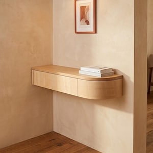 May include: A light-toned wooden floating wall shelf featuring a rectangular drawer section and a curved end piece. Two white books are placed on the flat top surface of the shelf, which is mounted against a beige wall below a small framed print.