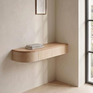 Peut inclure: Une étagère murale flottante en bois clair avec un bord gauche arrondi et une façade de tiroir rectangulaire. Deux livres fins sont empilés sur la surface supérieure de l'étagère. L'étagère est fixée sur un mur uni clair près d'une fenêtre.
