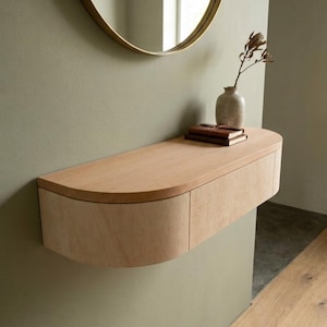 May include: A wall-mounted floating console table crafted from light-toned wood, featuring a semi-circular top and a curved front panel with two integrated drawers. The surface holds a small textured ceramic vase with a dried stem and two stacked books.
