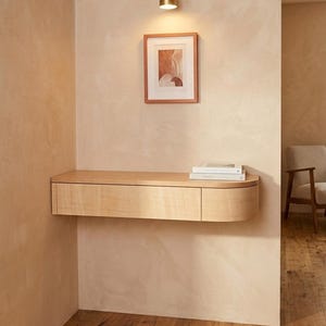 May include: A light-toned wooden floating wall-mounted console table with a single drawer and a rounded right-side edge. The console features a smooth, flat top surface and is positioned against a textured beige wall beneath a framed print.