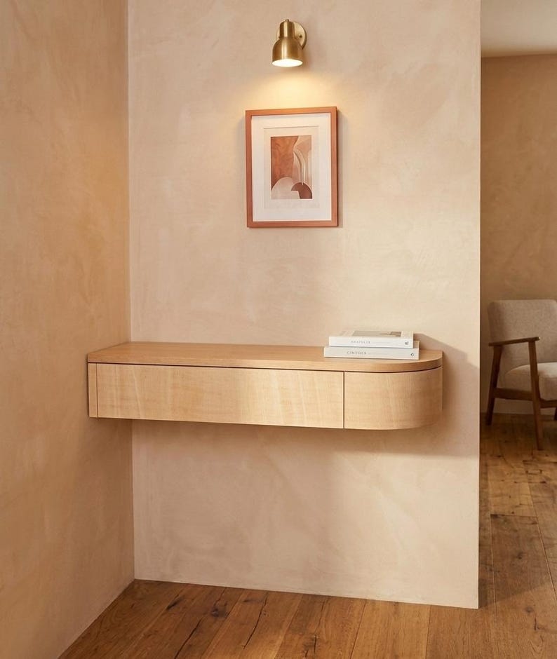 May include: A light-toned wooden floating wall-mounted console table with a single drawer and a rounded right-side edge. The console features a smooth, flat top surface and is positioned against a textured beige wall beneath a framed print.