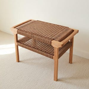 May include: A two-tiered bench with a woven top and shelf. The frame is made of light-colored wood, and the woven surfaces are a warm brown. The bench has four sturdy legs and a rectangular shape, suitable for indoor use.
