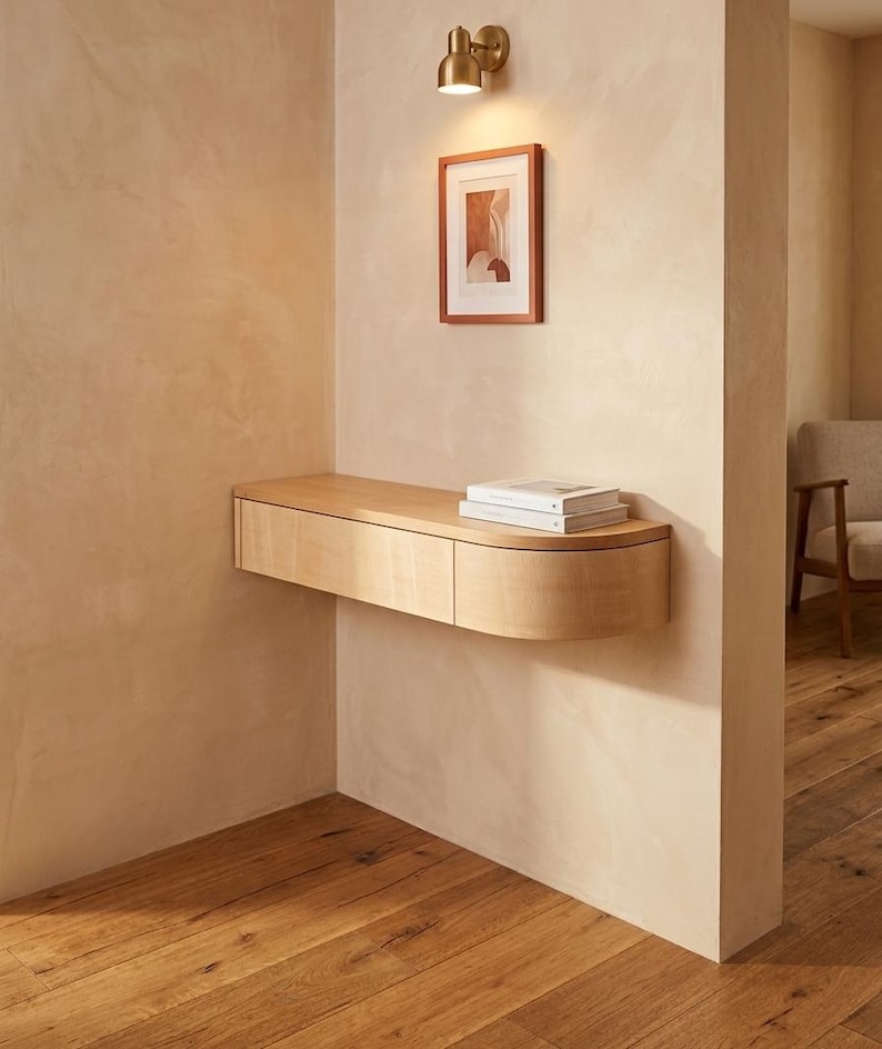 May include: A light-toned wooden floating wall shelf featuring a rectangular drawer section and a curved end piece. Two white books are placed on the flat top surface of the shelf, which is mounted against a beige wall below a small framed print.