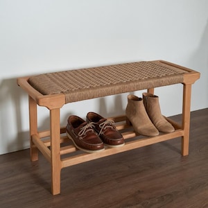 Könnte beinhalten: Schuhbank aus Holz mit gewebter Oberseite und unterem Regal. Die Bank ist aus hellem Holz und hat eine rechteckige Form. Braune Lederschuhe und hellbraune Wildlederstiefel sind auf der Bank platziert.