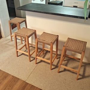 Op de afbeelding: Vier houten krukken met gevlochten zittingen. De krukken zijn in een rij op een lichtbruin tapijt geplaatst.