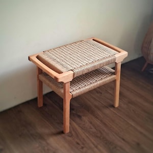 Könnte beinhalten: Kleiner, zweistufiger Holzschemel mit gewebter Sitzfläche und Ablage. Der Rahmen besteht aus hellem Holz, und die gewebten Oberflächen sind naturfarben beige. Der Hocker hat eine rechteckige Form und ist für verschiedene Zwecke geeignet.