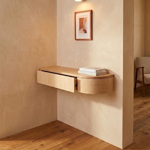 May include: A light-colored wooden floating wall-mounted console table with a single rectangular drawer and a rounded side edge. Two books are stacked on the flat top surface of the console, which is attached to a beige wall below a wall lamp.