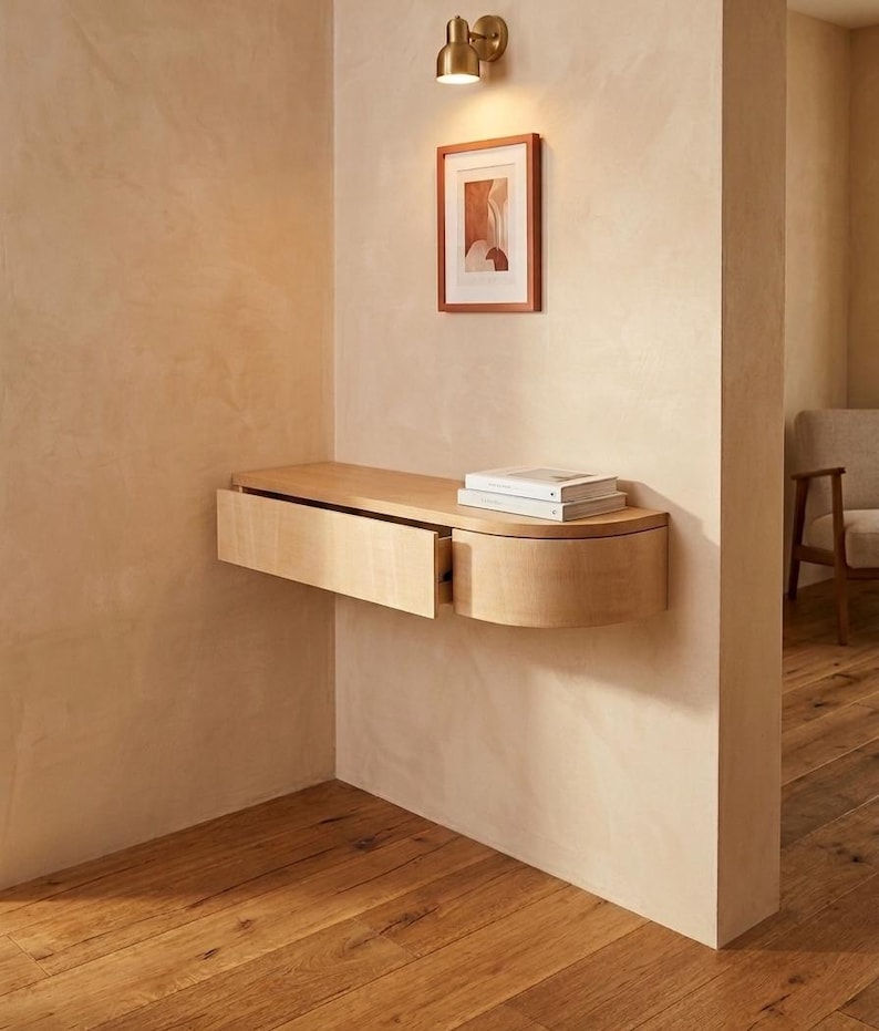 May include: A light-colored wooden floating wall-mounted console table with a single rectangular drawer and a rounded side edge. Two books are stacked on the flat top surface of the console, which is attached to a beige wall below a wall lamp.