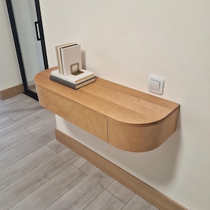 以下が含まれることがあります： 曲線的な前面と2つの引き出しを備えた、明るい色の木製フローティングシェルフ。シェルフは白い壁に取り付けられ、本と金色のブックエンドの山を保持しています。シェルフの長さは約91cmです。