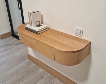 May include: A light-colored wooden floating shelf with a curved front and two drawers. The shelf is mounted on a white wall and holds a stack of books and a gold-colored bookend. The shelf is approximately 36 inches long.