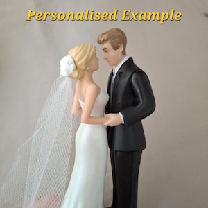 Pode incluir: Um topo de bolo de casamento com duas figuras frente a frente. Uma figura usa um vestido branco sem alças com um véu a condizer, e a outra um fato preto com uma camisa branca. O texto "Personalised Example" é visível acima deles.
