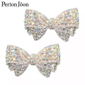 May include: Two white bows with iridescent rhinestones. The bows are made of a white fabric and are decorated with clear and iridescent rhinestones.