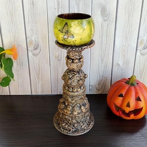 May include: A decorative candle holder with a green bowl-shaped top featuring a butterfly design. The holder is supported by a textured, brown, ornate pedestal. An orange pumpkin with a carved face sits nearby.