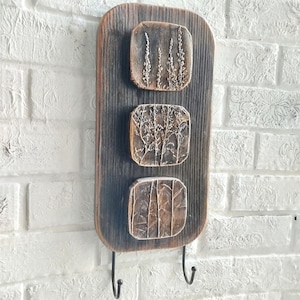 May include: A rustic wooden wall-mounted key holder with three square, textured panels featuring botanical designs. The dark brown wood has a distressed finish, and two metal hooks are attached at the bottom. The background is a white brick wall.