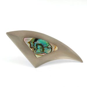 May include: A curved, fan-shaped brooch made of brushed silver-toned metal. The centre features an inlaid triangular piece of iridescent abalone shell displaying swirling patterns of green, blue, and pearlescent white colours.