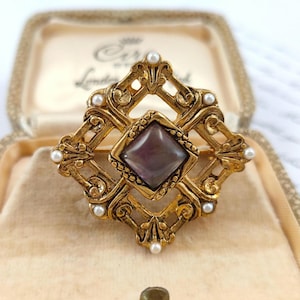 May include: A vintage gold-tone brooch with a central square, dark purple stone. The brooch features an ornate, openwork design with small, round, white pearls. The brooch is in a beige box.