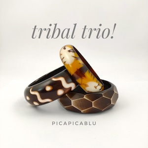 May include: Three bangle bracelets with the text tribal trio! and PICAPICABLU. One is dark brown with white wavy lines, one is amber with cream patterns, and one is dark brown with a faceted hexagonal surface texture.