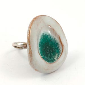 May include: A ring featuring a large, irregular, teardrop-shaped focal piece with a glossy white finish and a textured, deep emerald green center. The edges of the focal piece show a thin, rustic copper-colored rim, attached to a simple silver band.