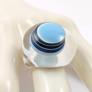 May include: A clear, chunky resin ring featuring a circular top inlay. The inlay displays a layered design with a light blue centre, followed by a thin white band, a black stripe, and a dark blue base, creating a stacked, multi-coloured effect.