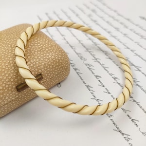 Peut inclure: Bracelet jonc crème avec un motif de corde torsadée. Le bracelet présente un accent en spirale marron clair. Le bijou est présenté sur une surface texturée de couleur beige, avec une boîte de style vintage en arrière-plan.