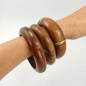 May include: Three thick wooden bangle bracelets with a polished finish. One bracelet features inlaid gold-tone metal leaf-shaped accents, while another includes a smooth gold-tone metal band detail. The wood displays natural grain patterns.