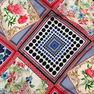 Op de afbeelding: Een stof met een patchworkontwerp van vierkante panelen. De patronen omvatten donkerblauwe stippen, roze pioenen met gouden krulwerk, blauwe klaprozen en veelkleurige bloemstukken, allemaal gescheiden door rode en witte geometrische randen.