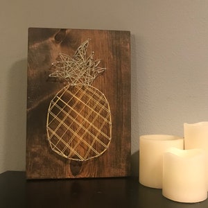 Op de afbeelding: Een houten bord met een string art ananas ontwerp. De ananas is gemaakt met gouden en zilveren draad. Het bord staat op een zwart oppervlak met drie witte kaarsen.
