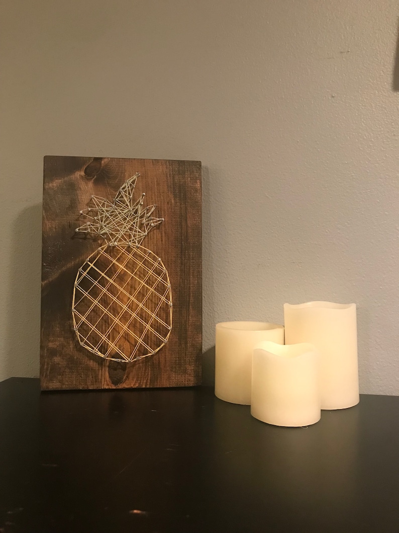 May include: A wooden sign with a string art pineapple design. The pineapple is made with gold and silver string. The sign is sitting on a black surface with three white candles.