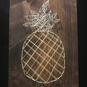 May include: A string art pineapple with a white top and a yellow body on a brown wooden background.