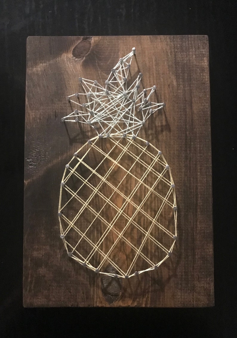 May include: A string art pineapple with a white top and a yellow body on a brown wooden background.