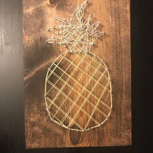 May include: A wooden board with a string art pineapple design. The pineapple is made with silver nails and yellow string.