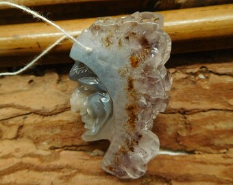 May include: A carved gemstone pendant in the shape of a Native American profile. The pendant is a mix of translucent lavender, gray, and brown hues, with a white string attached for wearing. The stone has a rough, textured surface.