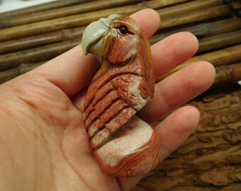 May include: Hand-carved parrot figurine made of red, white, and brown stone. The parrot is perched on a branch with its wings spread out.
