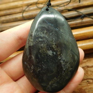 May include: A smooth, black stone pendant with a dark green and gray marbled pattern. The pendant is teardrop shaped and has a hole at the top for a cord.
