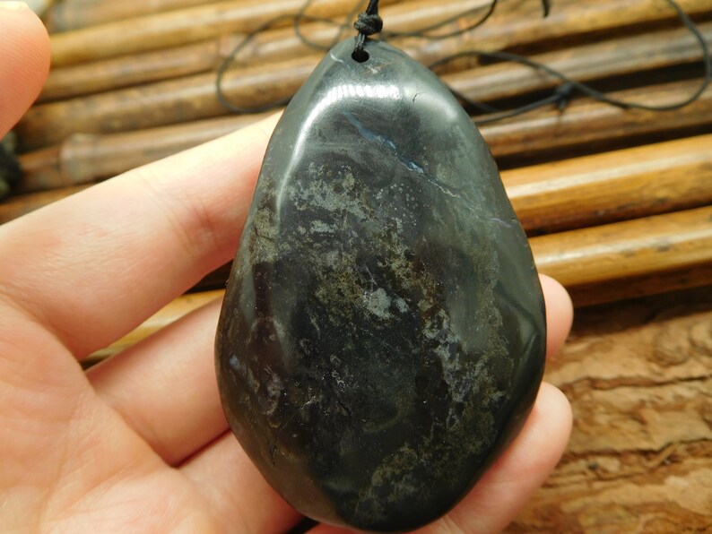 May include: A smooth, black stone pendant with a dark green and gray marbled pattern. The pendant is teardrop shaped and has a hole at the top for a cord.