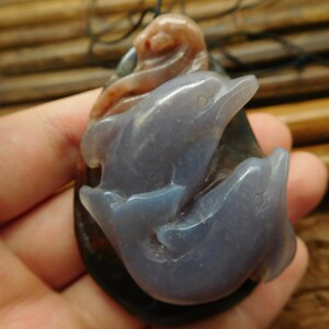 May include: A pendant featuring two carved dolphins made of purple agate. The dolphins are intertwined and have a natural, organic look. The pendant is attached to a black cord.