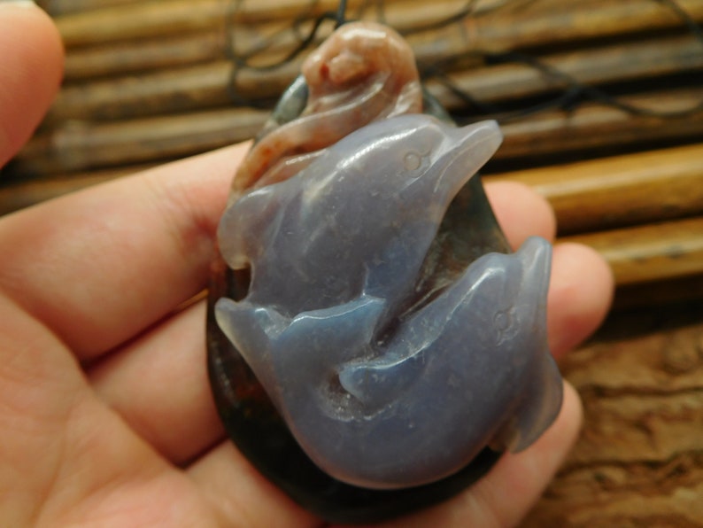 May include: A pendant featuring two carved dolphins made of purple agate. The dolphins are intertwined and have a natural, organic look. The pendant is attached to a black cord.