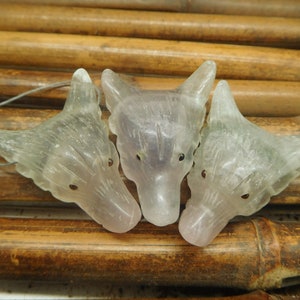 Puede incluir: Tres colgantes tallados en forma de cabeza de lobo hechos de amatista transparente y translúcida con un tono púrpura claro. Los colgantes están dispuestos en fila, con el colgante del medio ligeramente más alto que los otros dos.