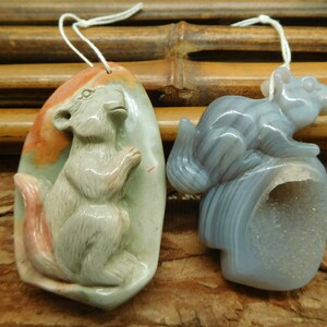 Puede incluir: Dos colgantes de ardilla tallados en piedra. Uno es una ardilla gris claro con una cola rosa tallada en una piedra multicolor. El otro es una ardilla gris tallada en una ágata azul claro con una geoda de cristal.