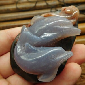 May include: A pendant carved from agate featuring three dolphins, one on top of the other. The dolphins are a light purple color and the base is a dark gray.