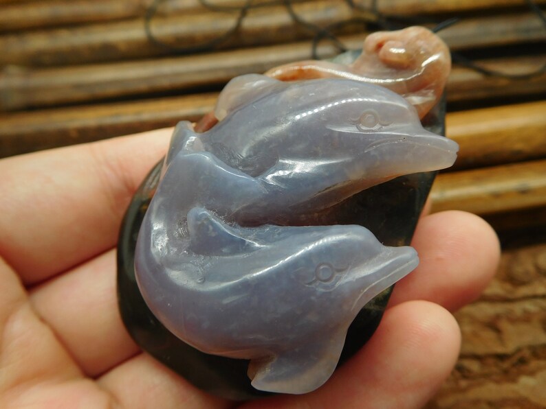 May include: A pendant carved from agate featuring three dolphins, one on top of the other. The dolphins are a light purple color and the base is a dark gray.
