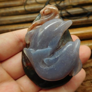 May include: A pendant carved from agate featuring two dolphins, one swimming above the other. The dolphins are a light blue color, while the agate is a mottled brown and black.