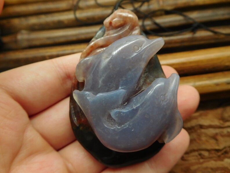 May include: A pendant carved from agate featuring two dolphins, one swimming above the other. The dolphins are a light blue color, while the agate is a mottled brown and black.