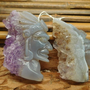 Puede incluir: Dos colgantes de piedra de amatista tallados, uno con una cara tallada en la piedra y el otro con un dise&ntilde;o m&aacute;s abstracto. Ambos colgantes tienen una textura rugosa y natural y son de color p&uacute;rpura claro.
