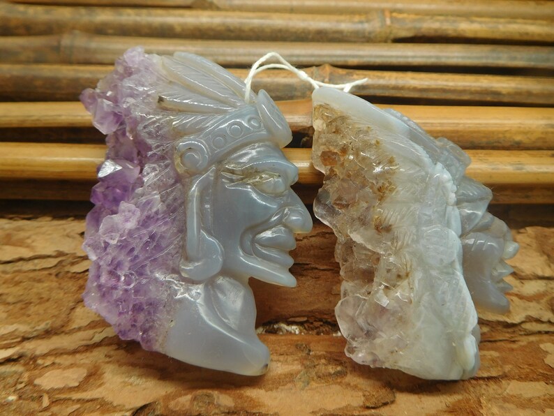 Puede incluir: Dos colgantes de piedra de amatista tallados, uno con una cara tallada en la piedra y el otro con un dise&ntilde;o m&aacute;s abstracto. Ambos colgantes tienen una textura rugosa y natural y son de color p&uacute;rpura claro.